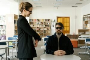 Two individuals interacting in a library, both wearing sunglasses, suggesting a context of visual impairment or fashion.